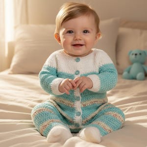 May include: A baby in a handmade crochet romper in white, light blue, and beige. The romper features a button-up front and long sleeves. A light blue teddy bear is on the bed in the background.