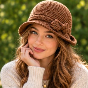 Könnte beinhalten: Ein einfarbiger, brauner Häkel-Bucket-Hat mit strukturiertem Strickmuster und einer kleinen, passenden Häkelschleife an der Seite. Der Hut hat eine abgerundete Krone und eine leicht nach unten geneigte Krempe aus erdfarbenem Garn.