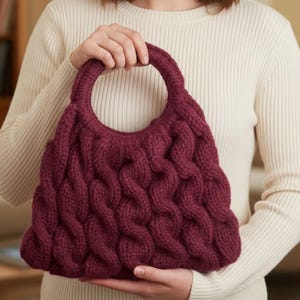 May include: A burgundy-colored, hand-knitted handbag with a large circular handle. The bag features a textured, cable-knit design. The bag is held by a person wearing a cream-colored sweater.