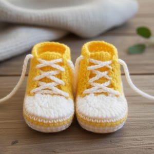 May include: A pair of handmade baby sneakers in bright yellow and white. The crochet shoes feature white laces and a classic sneaker design. The shoes are displayed on a wooden surface, with a soft, white fabric in the background.