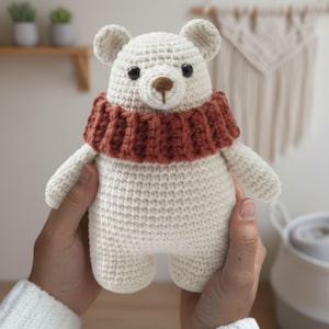 May include: A handmade, off-white crochet bear with a rust-colored scarf. The bear has small black eyes and a brown nose. The bear is being held by a person. The bear is a soft toy.
