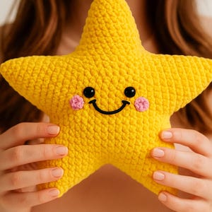 Könnte beinhalten: Gelber, gehäkelter Stern mit einem lächelnden Gesicht. Der Stern hat schwarze Knopfaugen und rosa Wangen. Das handgefertigte Spielzeug hat eine strukturierte Oberfläche. Ein fröhliches Design.