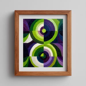 May include: Abstract geometric art print in a wooden frame. The artwork features a pattern of overlapping circles in shades of green, purple, and white against a dark blue background. The artwork is framed in a light brown wooden frame.