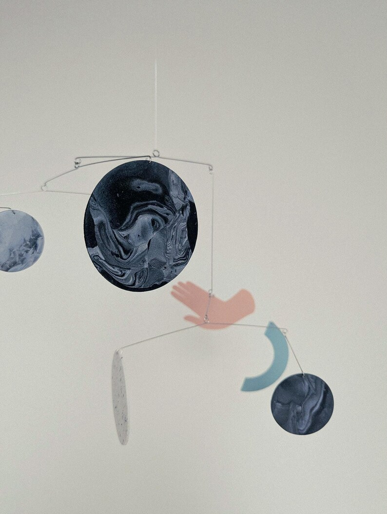 Peut inclure: Mobile moderne avec des formes circulaires et abstraites en bleu et blanc, suspendues par de fins fils. Une forme de main rose et un croissant bleu ajoutent des &eacute;l&eacute;ments uniques. Con&ccedil;u pour &ecirc;tre suspendu au plafond, c'est une &oelig;uvre d'art.
