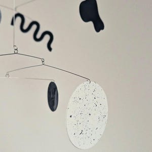 Peut inclure: Mobile sculpturale moderne au design minimaliste. Il pr&eacute;sente une s&eacute;rie de formes, dont un cercle blanc mouchet&eacute;, une ligne noire ondul&eacute;e et d'autres formes sombres, suspendues par de fines tiges m&eacute;talliques. Le mobile est con&ccedil;u pour &ecirc;tre suspendu et tourner.