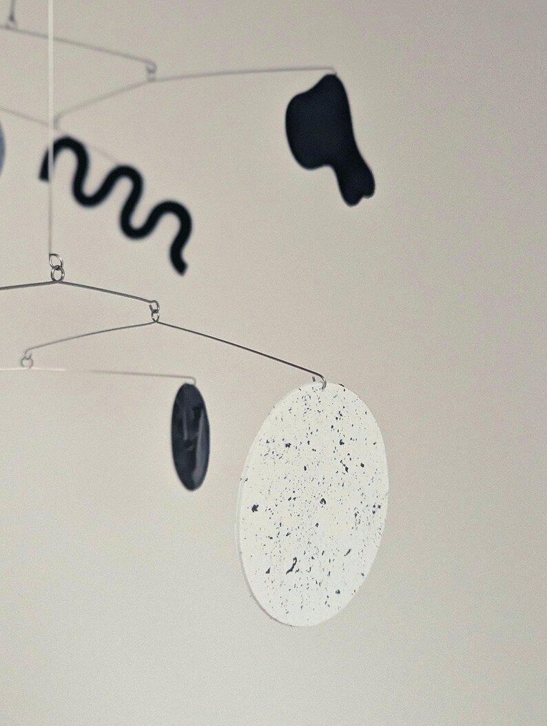Peut inclure: Mobile sculpturale moderne au design minimaliste. Il pr&eacute;sente une s&eacute;rie de formes, dont un cercle blanc mouchet&eacute;, une ligne noire ondul&eacute;e et d'autres formes sombres, suspendues par de fines tiges m&eacute;talliques. Le mobile est con&ccedil;u pour &ecirc;tre suspendu et tourner.