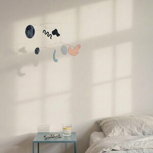 Peut inclure: Mobile moderne avec des formes abstraites noires, bleues et roses. Les formes comprennent une main, un cercle et une ligne ondul&eacute;e. Le mobile est suspendu &agrave; un fil fin contre un mur blanc. Une petite table bleue avec des lunettes et un verre d'eau est au premier plan.