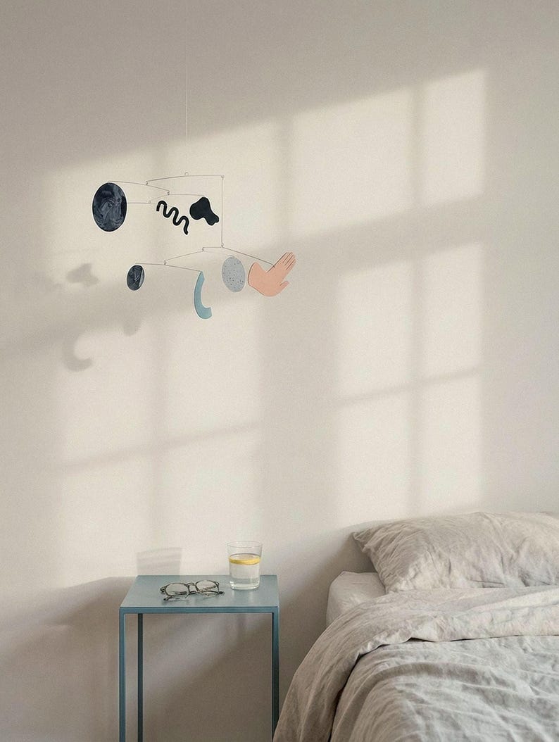 Peut inclure: Mobile moderne avec des formes abstraites noires, bleues et roses. Les formes comprennent une main, un cercle et une ligne ondul&eacute;e. Le mobile est suspendu &agrave; un fil fin contre un mur blanc. Une petite table bleue avec des lunettes et un verre d'eau est au premier plan.
