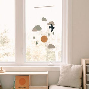 Op de afbeelding: Een decoratieve mobiel met een vogel, wolken, zon en regendruppels, hangend voor een raam. De mobiel heeft een natuurlijke, minimalistische uitstraling met zachte kleuren. Een kleine houten plank met het boek "abecadio" is ook zichtbaar.