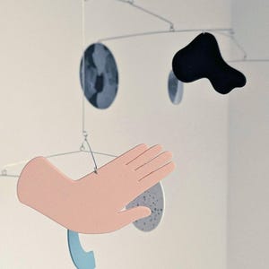 Peut inclure: Mobile artistique contemporain avec des formes vari&eacute;es. Il comprend une main rose clair, une forme noire abstraite, une forme bleue incurv&eacute;e et deux formes circulaires mouchet&eacute;es. L'ensemble est suspendu par des tiges m&eacute;talliques fines, cr&eacute;ant un &eacute;quilibre.