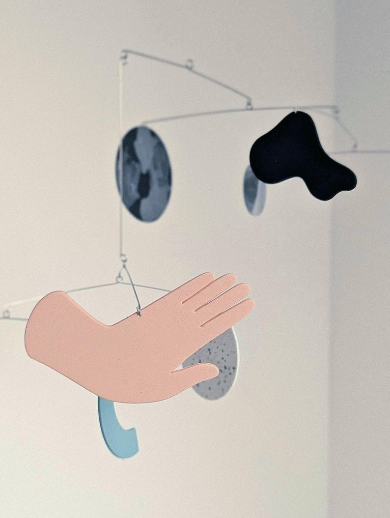 Peut inclure: Mobile artistique contemporain avec des formes vari&eacute;es. Il comprend une main rose clair, une forme noire abstraite, une forme bleue incurv&eacute;e et deux formes circulaires mouchet&eacute;es. L'ensemble est suspendu par des tiges m&eacute;talliques fines, cr&eacute;ant un &eacute;quilibre.