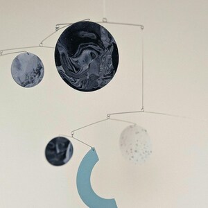 Peut inclure: Mobile moderne au design minimaliste. Il pr&eacute;sente des &eacute;l&eacute;ments circulaires et en forme de croissant dans des tons de bleu, gris et blanc, suspendus &agrave; de fines tiges m&eacute;talliques. Le plus grand cercle a un motif marbr&eacute;.
