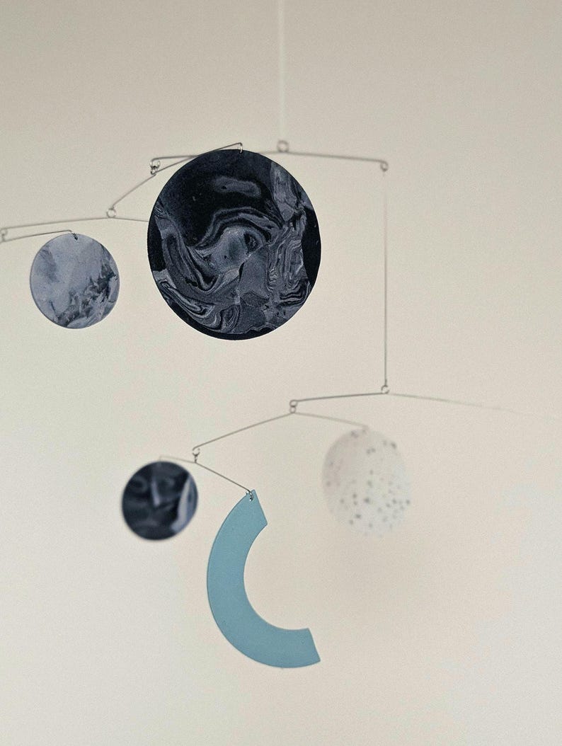 Peut inclure: Mobile moderne au design minimaliste. Il pr&eacute;sente des &eacute;l&eacute;ments circulaires et en forme de croissant dans des tons de bleu, gris et blanc, suspendus &agrave; de fines tiges m&eacute;talliques. Le plus grand cercle a un motif marbr&eacute;.