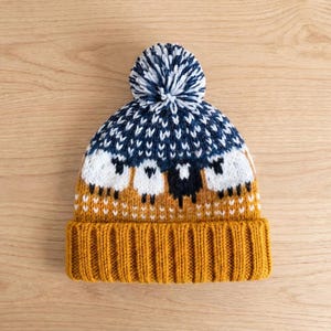 May include: A knitted beanie featuring a mustard yellow ribbed cuff and a pattern of white and black sheep across the centre. The upper section has a navy blue and white geometric design, topped with a matching navy blue and white pom-pom.