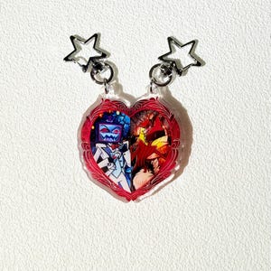 Op de afbeelding: Twee hartvormige sleutelhangers met stervormige sluitingen. Elke sleutelhanger heeft een kleurrijke illustratie van personages uit een show. E&eacute;n kant is blauw en wit, de andere is rood en oranje. De sleutelhangers zijn bevestigd aan zilveren metalen ringen.