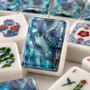 May include: A rectangular mahjong tile featuring a vibrant, iridescent abalone shell inlay with deep blue, teal, and purple tones, set within a smooth white base. This tile is surrounded by other mahjong tiles displaying various colourful designs.