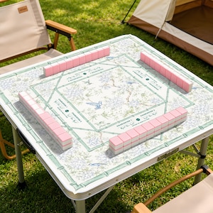 Könnte beinhalten: Ein quadratischer Klapptisch mit weißer Oberfläche, verziert mit einem grünen Bambusrahmenmuster, blauen Blumenmotiven und kleinen Vögeln. Vier Reihen rosa Mahjong-Steine liegen an den Rändern, mit dem Text Charleston 1, 2 und 3.