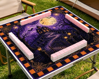 May include: A square mahjong table mat featuring a purple and black Halloween-themed illustration of a haunted house, a full moon, and spider webs. The mat is bordered by an orange and black checkered pattern and includes light pink mahjong tiles.