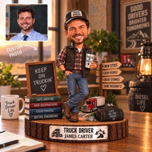 May include: A custom bobblehead figurine of a truck driver wearing a plaid shirt, vest, jeans, and a cap, standing on a wooden base. The display includes a chalkboard sign, a stack of books, a miniature red truck, and a signpost with road-themed text.