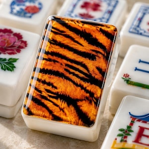 May include: A rectangular mahjong tile featuring a glossy, orange and black tiger stripe pattern on its front face, surrounded by other white mahjong tiles decorated with various colorful floral and traditional graphic designs.