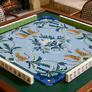 May include: A square mahjong table mat featuring a light blue surface with a repeating pattern of leopards, palm leaves, and a central floral motif. The mat is surrounded by white mahjong tiles with black and red characters and green backs.