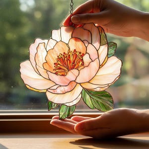 Puede incluir: Una peonía de vidriera en tonos melocotón, rosa y verde. La flor está suspendida por una cadena y presenta pétalos y hojas detallados. La luz atraviesa el cristal, creando una hermosa exhibición.