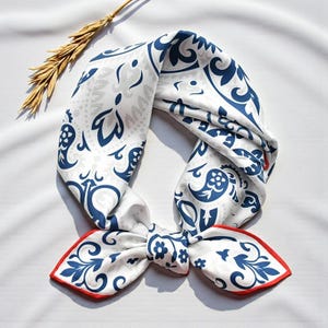 May include: White and blue patterned scarf with a red border. The scarf features a repeating floral and paisley design. A sprig of wheat is placed near the scarf.