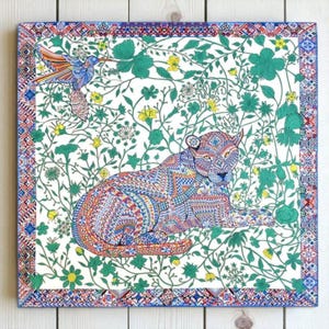 May include: Square artwork depicting a patterned lion resting amongst a floral and vine background. A hummingbird is shown in flight. The border has a geometric design with blue, red, and white accents.