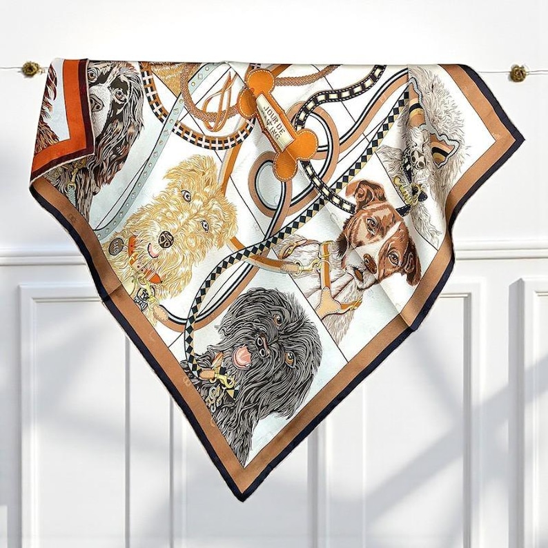 May include: Square silk scarf featuring a dog-themed design. The scarf has a white background with illustrations of various dog breeds, along with dog collars and leashes. The border is brown with a navy blue trim. The text "JOUR DE F&Ecirc;TE" is on a bone-shaped tag.