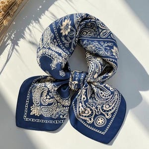 May include: A square silk scarf in deep navy blue featuring a cream-colored paisley and floral pattern. The fabric has a smooth, satin-like finish with a decorative border design. The scarf is tied in a knot, displaying its intricate print.