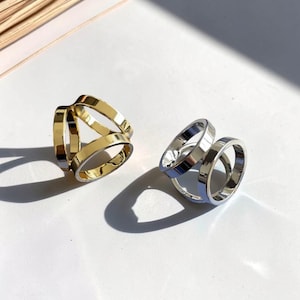 May include: Two rings, one gold-toned and one silver-toned, are displayed on a white surface. Each ring features a unique design with multiple bands, creating an open, geometric shape. The rings are positioned to catch the light, highlighting their metallic sheen.
