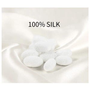 May include: A collection of white silk cocoons, with a textured surface, are arranged on a white, silky fabric. The text "100% SILK" is displayed above the cocoons. The image has a soft, bright appearance.