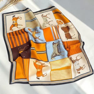 May include: A square silk scarf featuring a grid pattern of orange, light blue, and cream panels. The design includes illustrations of horses wearing blankets, striped sections, and a black border. The scarf has a smooth, reflective finish.