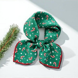 May include: A green Christmas scarf with a red border, adorned with white stars and candy cane patterns. The scarf is tied in a bow shape, perfect for holiday wear. A sprig of evergreen is in the background.