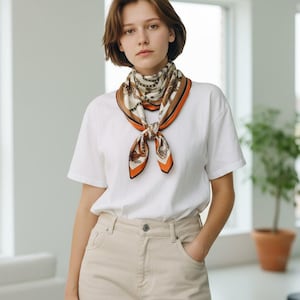 May include: A silk scarf with a cream background and a brown and orange border is tied around the neck. The scarf features a repeating pattern of ornate designs. The person is wearing a white t-shirt and tan pants.