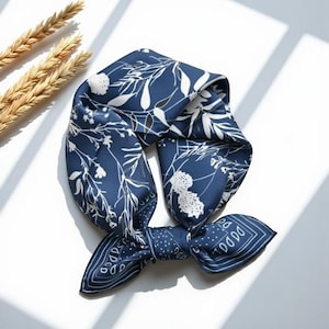 May include: A navy blue silk scarf with a white floral pattern. The scarf is square-shaped and tied in a knot. The scarf is lying on a white surface with a few stalks of wheat in the upper left corner.