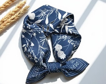 May include: A navy blue silk scarf with a white floral pattern. The scarf is square-shaped and tied in a knot. The scarf is lying on a white surface with a few stalks of wheat in the upper left corner.
