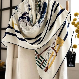 May include: Cream-colored silk scarf with a navy blue and white striped border. The scarf features equestrian-themed illustrations in shades of blue, green, and brown. The scarf is draped over a white coat.