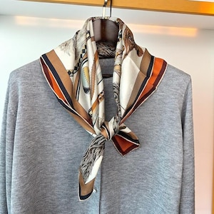 May include: A square silk scarf with a detailed equestrian-themed design in shades of cream, brown, and orange. The scarf is knotted and draped over a gray sweater on a wooden hanger, showcasing its intricate patterns and colors.