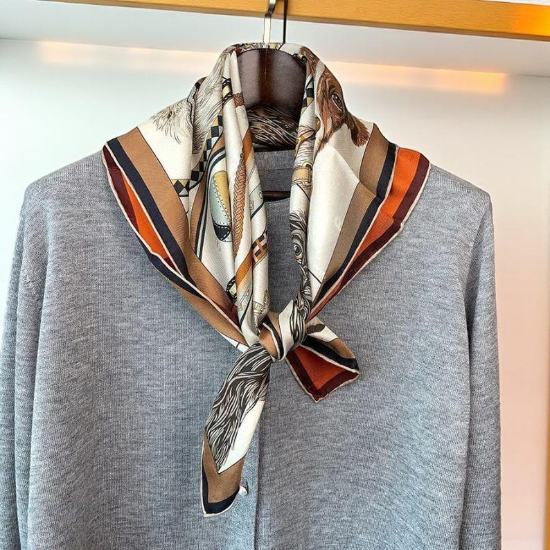 May include: A square silk scarf with a detailed equestrian-themed design in shades of cream, brown, and orange. The scarf is knotted and draped over a gray sweater on a wooden hanger, showcasing its intricate patterns and colors.