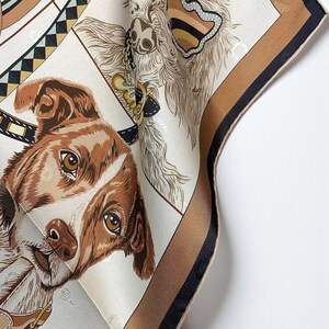 May include: A silk scarf featuring detailed illustrations of dogs. The scarf has a cream background with brown and black borders. The dogs are depicted with brown and white fur, and the scarf is folded to show multiple angles.