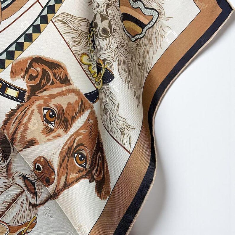 May include: A silk scarf featuring detailed illustrations of dogs. The scarf has a cream background with brown and black borders. The dogs are depicted with brown and white fur, and the scarf is folded to show multiple angles.
