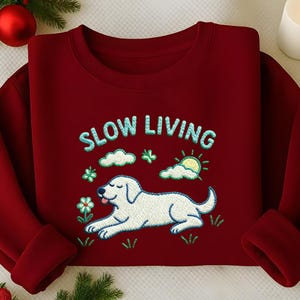 May include: A maroon sweatshirt with the embroidered phrase "SLOW LIVING" above a white dog illustration. The dog is lying down with its tongue out, surrounded by embroidered clouds, flowers, and a sun. The sweatshirt has a crew neck and long sleeves.
