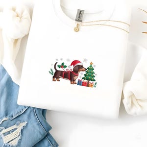 May include: White sweatshirt featuring a Christmas-themed dachshund wearing a Santa hat and a red harness. The design includes a decorated Christmas tree, wrapped gifts, and holly. A gold necklace is draped on the shirt.