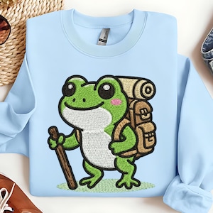 May include: Light blue sweatshirt featuring an embroidered cartoon frog with a backpack and walking stick. The frog is green with a white belly and pink cheeks. The backpack is brown, and the walking stick is brown.