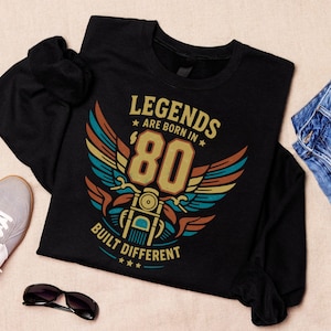 Op de afbeelding: Een zwart shirt met lange mouwen en een grafische print met de tekst 'LEGENDS ARE BORN IN 80 BUILT DIFFERENT' in geel, oranje en turquoise. Het ontwerp bevat een gestileerde motorfiets met vleugels en sterren in een retro kleurenpalet.