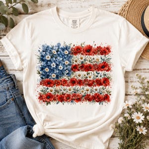 May include: A cream-colored short-sleeve t-shirt featuring a graphic print of the American flag composed entirely of blue hydrangeas, white daisies, and red poppies. The design is centered on the chest, showcasing a floral arrangement in the shape of the flag.
