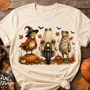 May include: A cream-colored t-shirt featuring a graphic of three frogs dressed for Halloween. One frog wears a witch hat and cape on a pumpkin, one rides a motorcycle while wearing a ghost sheet, and one is wrapped like a mummy among autumn leaves.