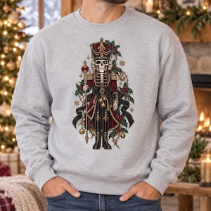May include: A light gray crewneck sweatshirt featuring a graphic print of a skeleton dressed as a nutcracker. The figure wears a red and black uniform with gold accents, holding a staff, surrounded by holly leaves, red berries, and gold bells.