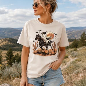 May include: A cream-colored short-sleeve t-shirt featuring a graphic print of a ghost wearing a cowboy hat riding a black horse. The design includes orange pumpkins, cacti, a large full moon, and small black bats against a desert landscape.